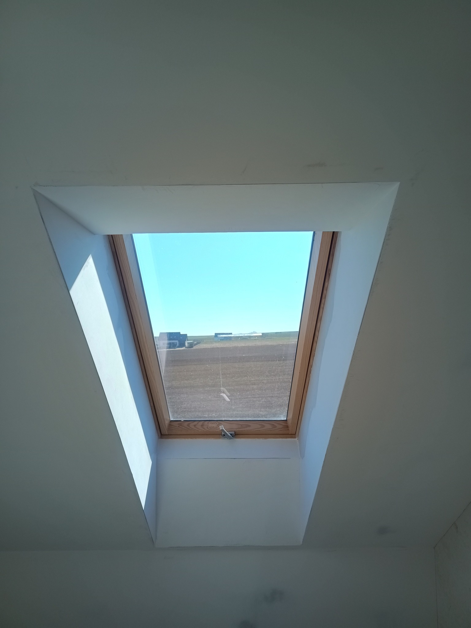 Wykończone wnęki okna dachowego na poddaszu, z widokiem na błękitne niebo i krajobraz za oknem. Świeżo pomalowane na biało ściany i drewniana rama okienna.