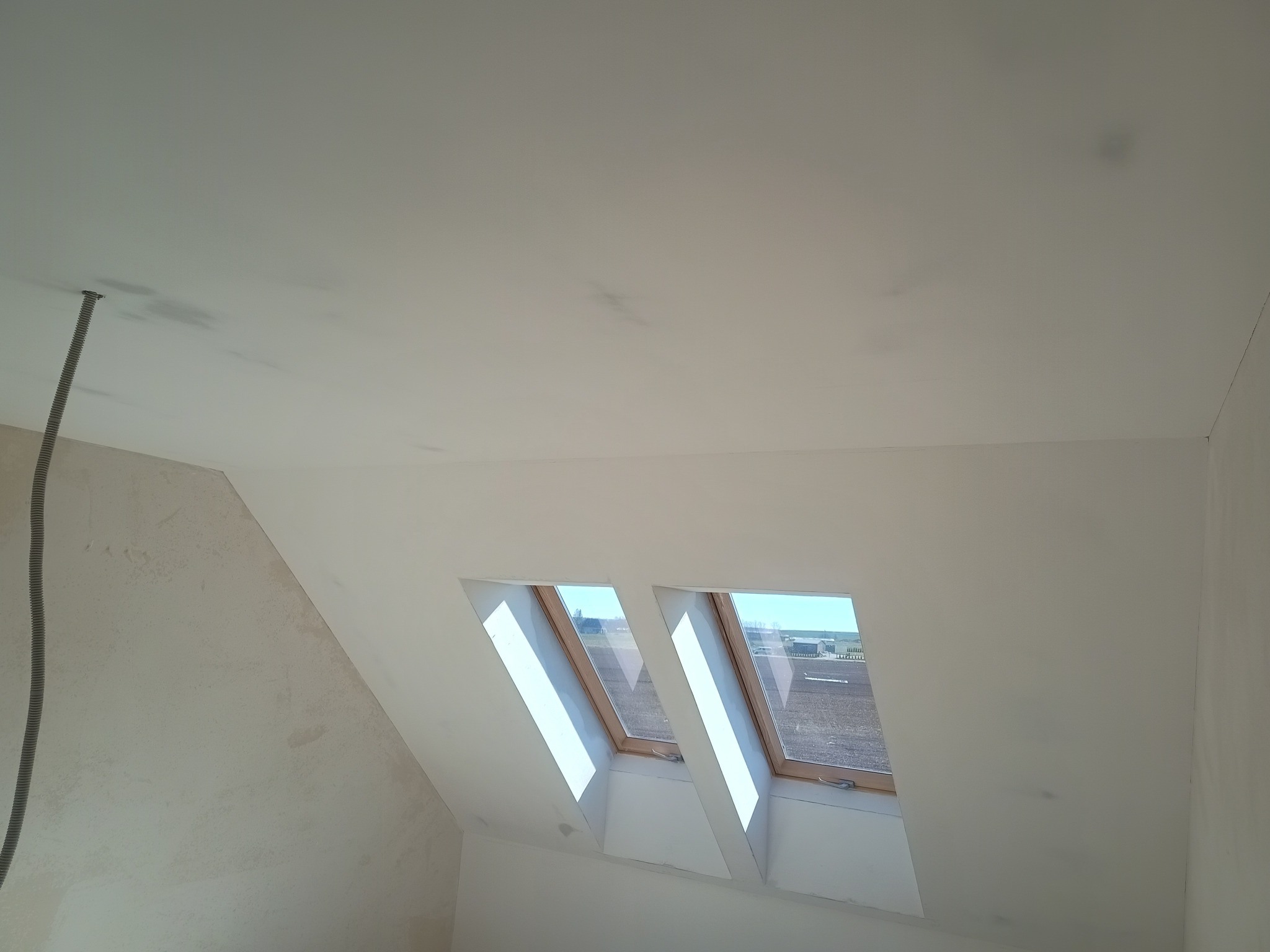 Białe, wyszpachlowane poddasze z dwoma oknami dachowymi, przez które wpada światło. Widoczne ślady po szpachlowaniu na ścianach i suficie. Kabel elektryczny w rogu.