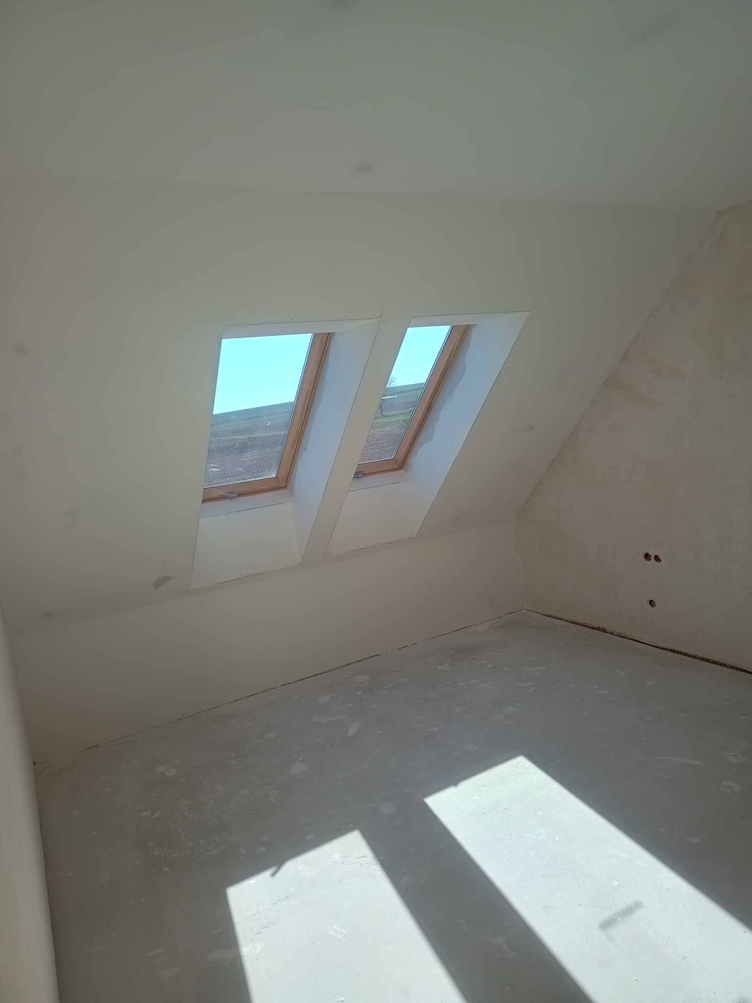 Poddasze po gładziowaniu, widoczne dwa okna dachowe i fragment ściany z gładzią. Podłoga przygotowana do dalszych prac wykończeniowych. Świeże, jasne wnętrze.