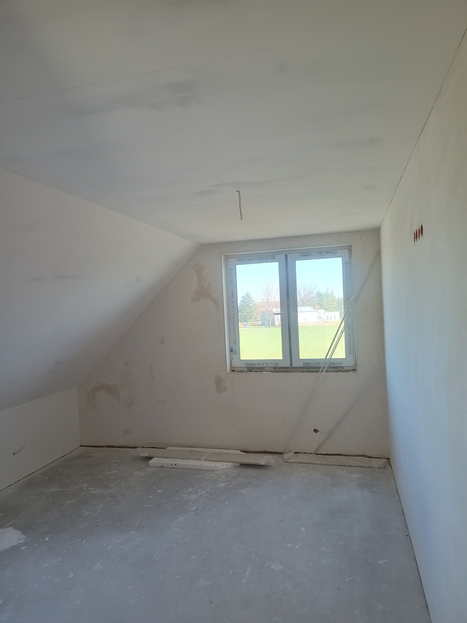 Pokój na poddaszu po szpachlowaniu, ściany przygotowane do malowania. Widoczne okno z widokiem na zewnątrz i fragment instalacji elektrycznej.