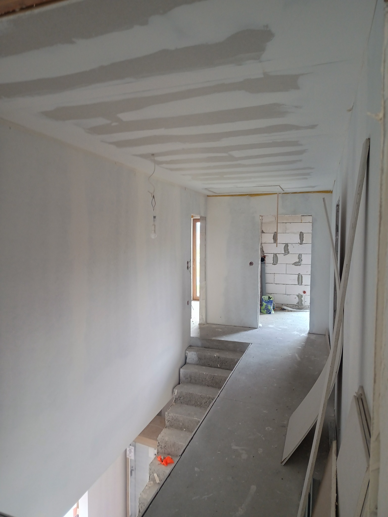 Korytarz w trakcie prac wykończeniowych: ściany i sufit po szpachlowaniu, przygotowane do malowania, widoczne płyty gipsowe i schody betonowe. Surowy stan.