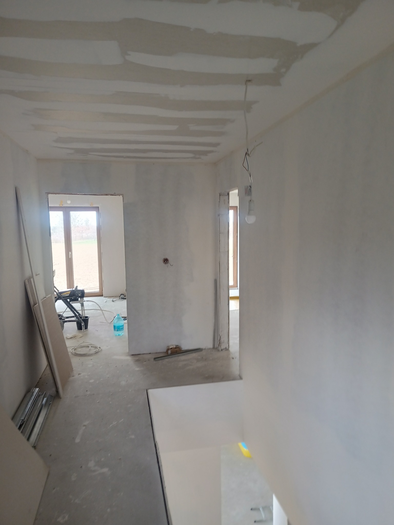 Wnętrze w trakcie prac wykończeniowych: surowy sufit po szpachlowaniu, ściany przygotowane do malowania, widoczny sprzęt malarski i płyty gipsowe.