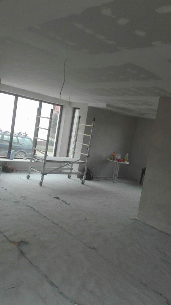 Wnętrze w trakcie szpachlowania, widoczne ślady po szpachli na suficie, rusztowanie na kółkach, okno z widokiem na samochód.