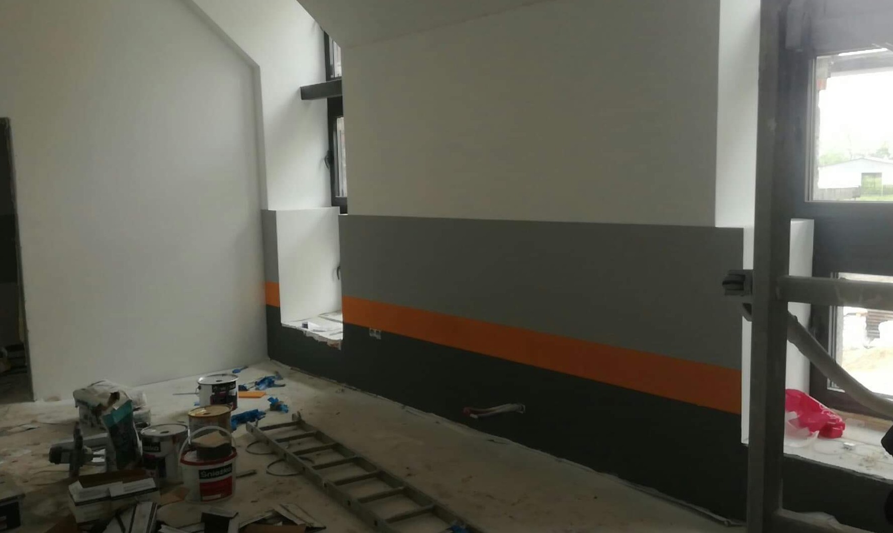Wnętrze w Przeworno w trakcie malowania: ściany z poziomymi pasami w odcieniach szarości i pomarańczy, widoczne puszki z farbą, drabina i narzędzia malarskie.