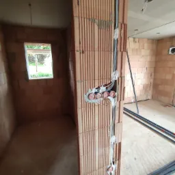 Wnętrze w stanie surowym z widoczną instalacją elektryczną w ścianie z cegły, kable prowadzone w peszlach, puszki elektryczne zamontowane w ścianie.