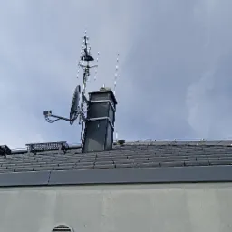 Anteny i talerz satelitarny zamontowane na kominie na dachu domu z szarej dachówki, widok z dołu.