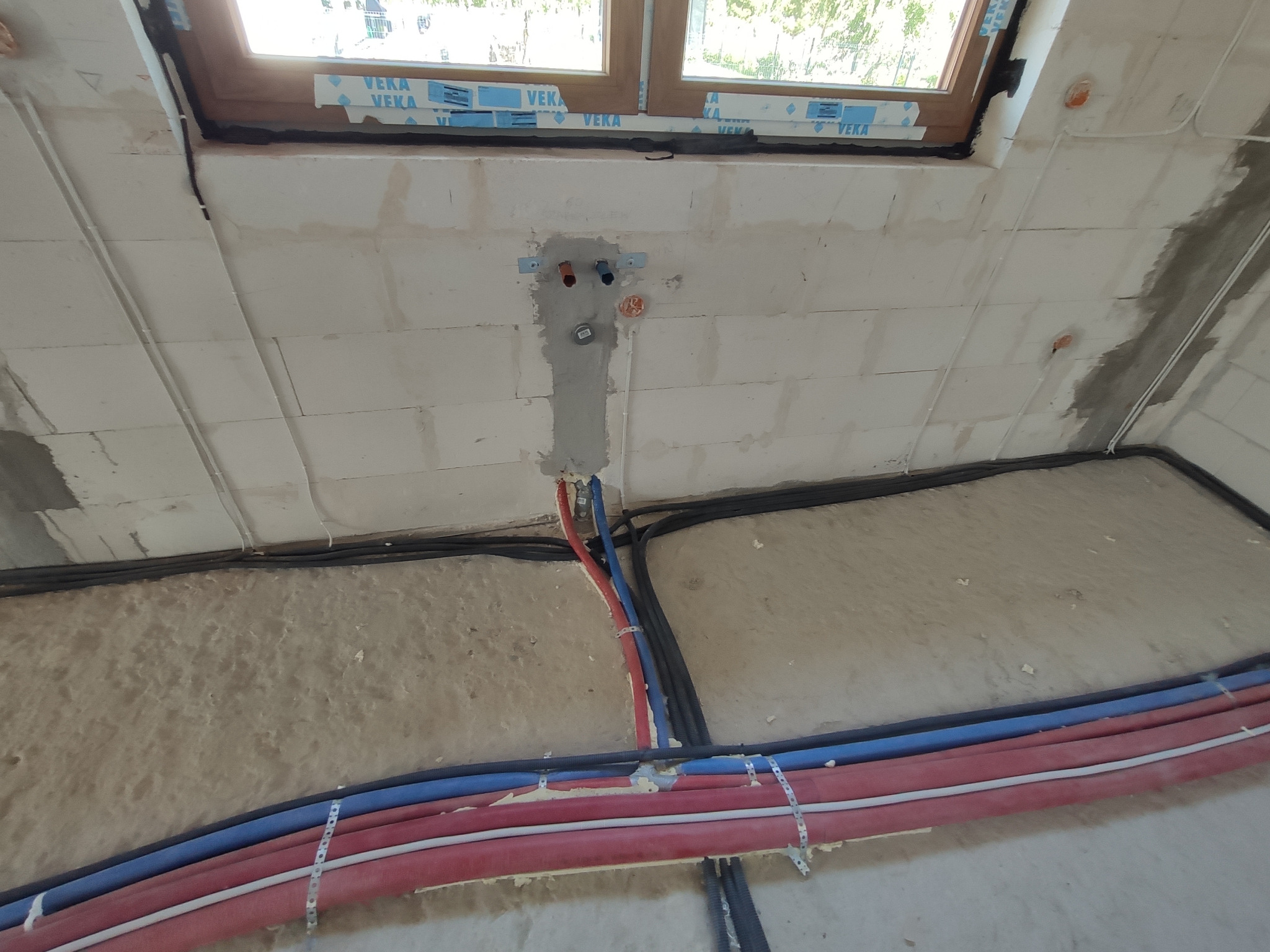 Surowy stan budynku w trakcie prac: widoczne rury instalacyjne w kolorach czerwonym i niebieskim, prowadzone wzdłuż ściany pod oknem, oraz czarne kable elektryczne, wszystko przymocowane metalowymi...