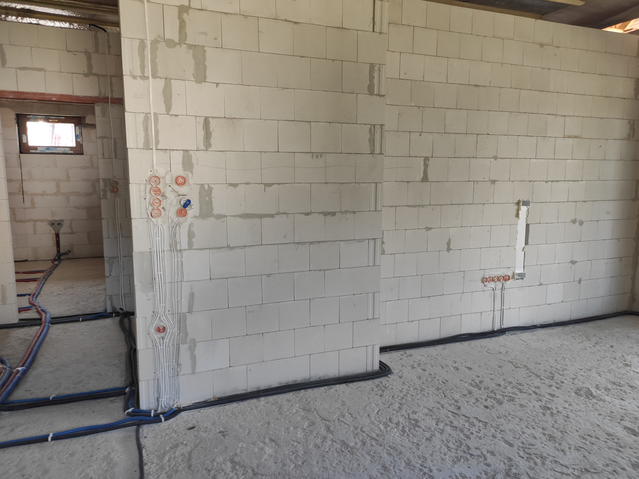 Surowy stan budynku z widocznymi przewodami elektrycznymi w ścianach z betonu komórkowego i rurami instalacyjnymi na podłodze.