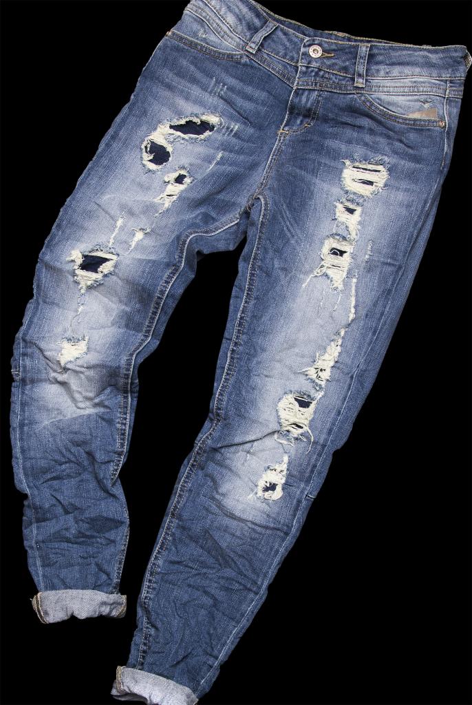 Stylowe, podarte jeansy damskie w odcieniach niebieskiego, z widocznymi przetarciami i dziurami, na czarnym tle. Nogawki podwinięte, ukazujące wewnętrzną stronę materiału.