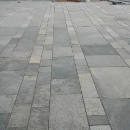 Ułożona z szarych płyt i kostki betonowej nawierzchnia tarasu, z zielonym krzewem i fragmentem schodów w tle.