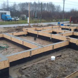 Wylane fundamenty z szalunkiem z płyty OSB i hydroizolacją pionową, widoczne zbrojenia i zalane betonem, na zewnątrz błoto i sprzęt budowlany.