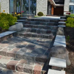 Kamienne schody prowadzące do domu, z elementami dekoracyjnymi z jasnego kamienia i otoczone roślinnością.