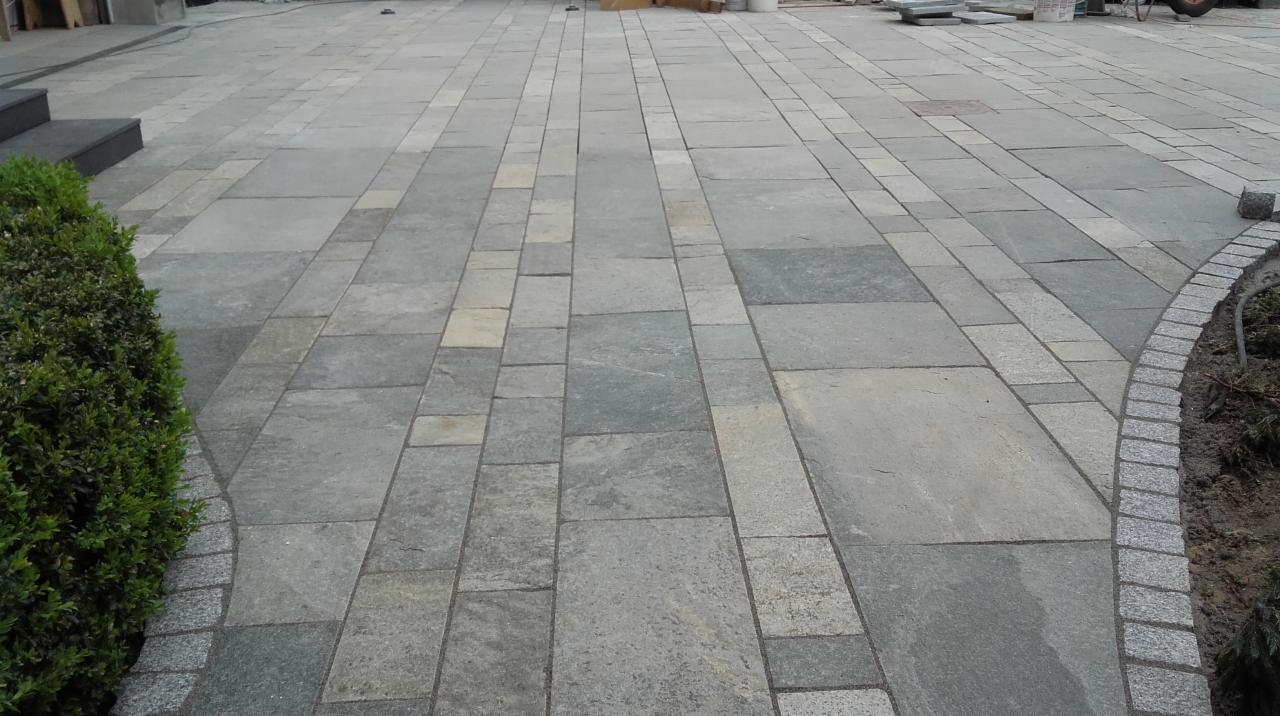 Ułożona z szarych płyt i kostki betonowej nawierzchnia tarasu, z zielonym krzewem i fragmentem schodów w tle.