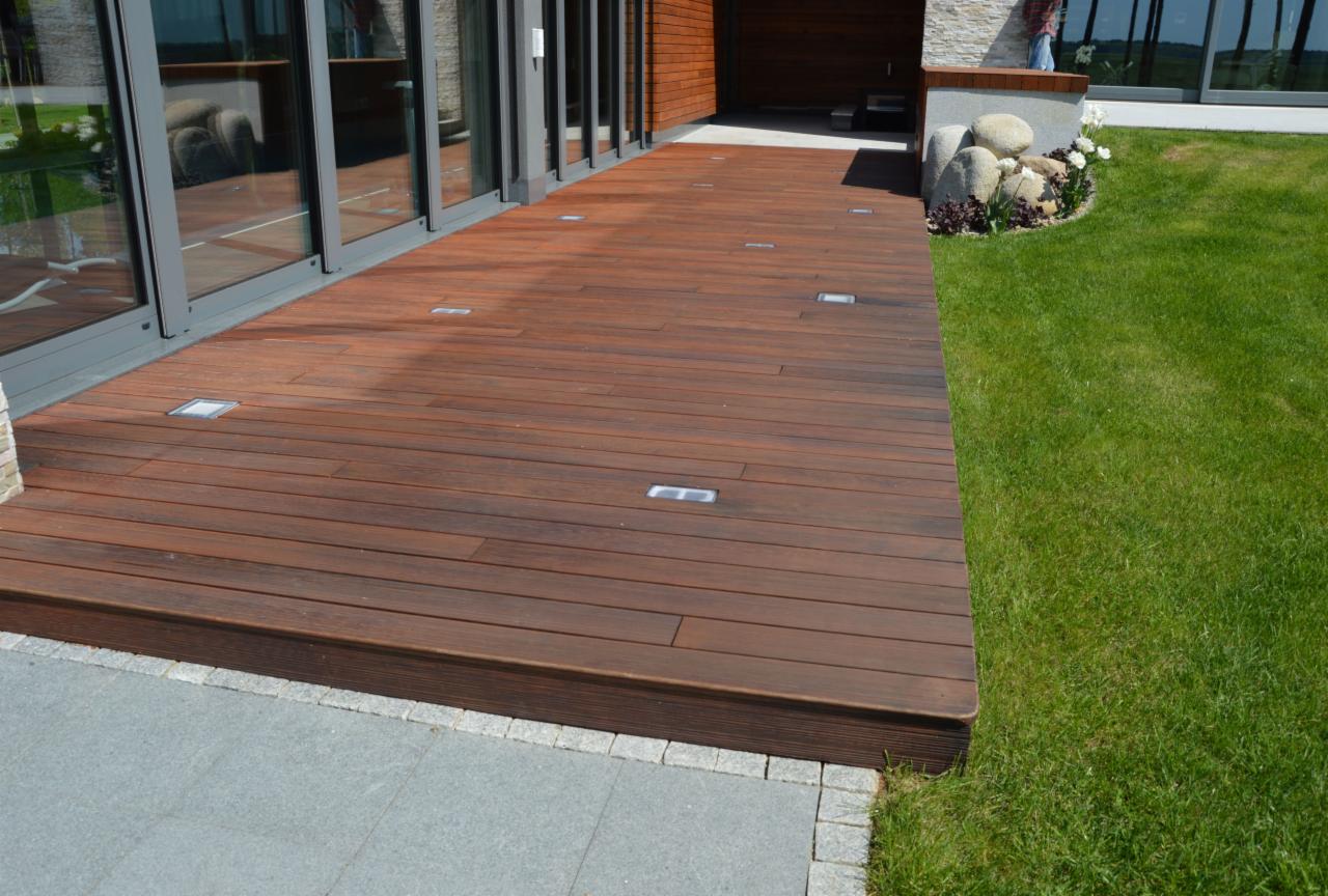 Nowoczesny taras z desek kompozytowych w kolorze ciemnego drewna, z wbudowanymi punktami świetlnymi, przylegający do budynku z dużymi oknami i zadbanym trawnikiem.