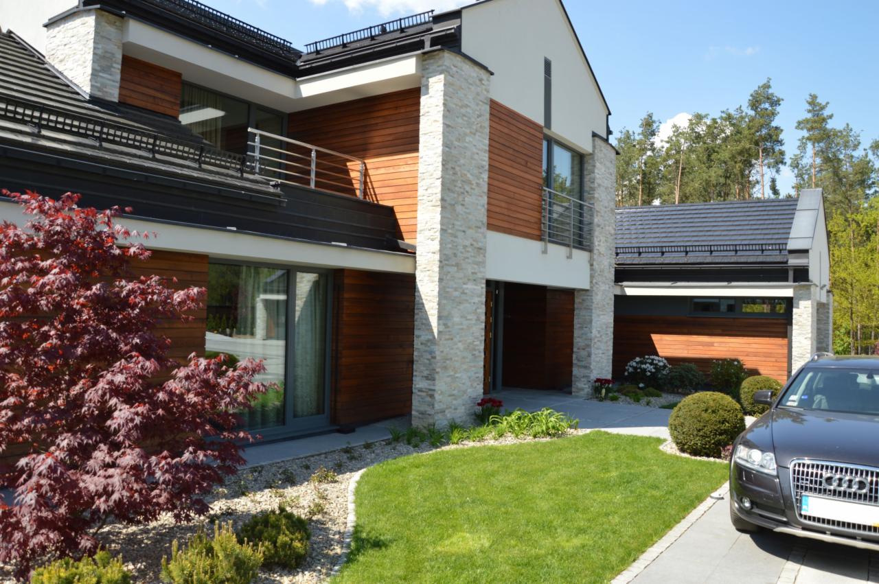 Nowoczesny dom z elewacją z drewna i kamienia, ciemna dachówka, zadbany trawnik i czerwony klon palmowy na pierwszym planie, szary samochód zaparkowany przed garażem.