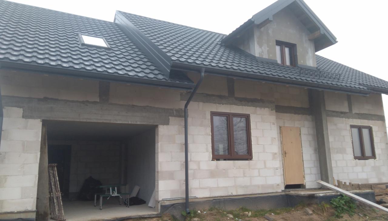 Dom murowany w stanie surowym zamkniętym z szarą blachodachówką, widoczne okna z brązowymi ramami i garaż z metalowymi rusztowaniami w środku.