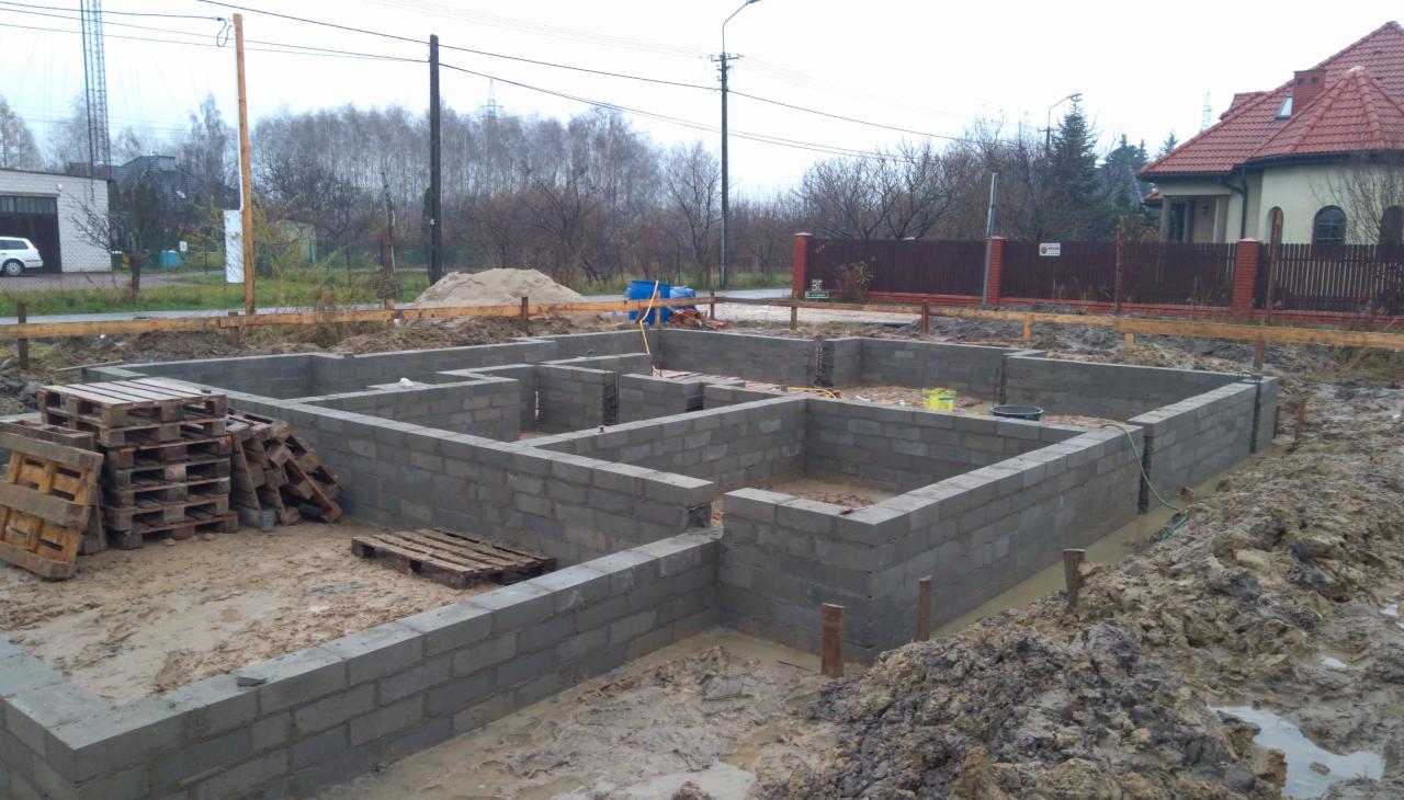 Fundamenty domu jednorodzinnego w trakcie budowy, bloczki betonowe ułożone w ziemi, widoczne drewniane palety i teren budowy w pochmurny dzień.