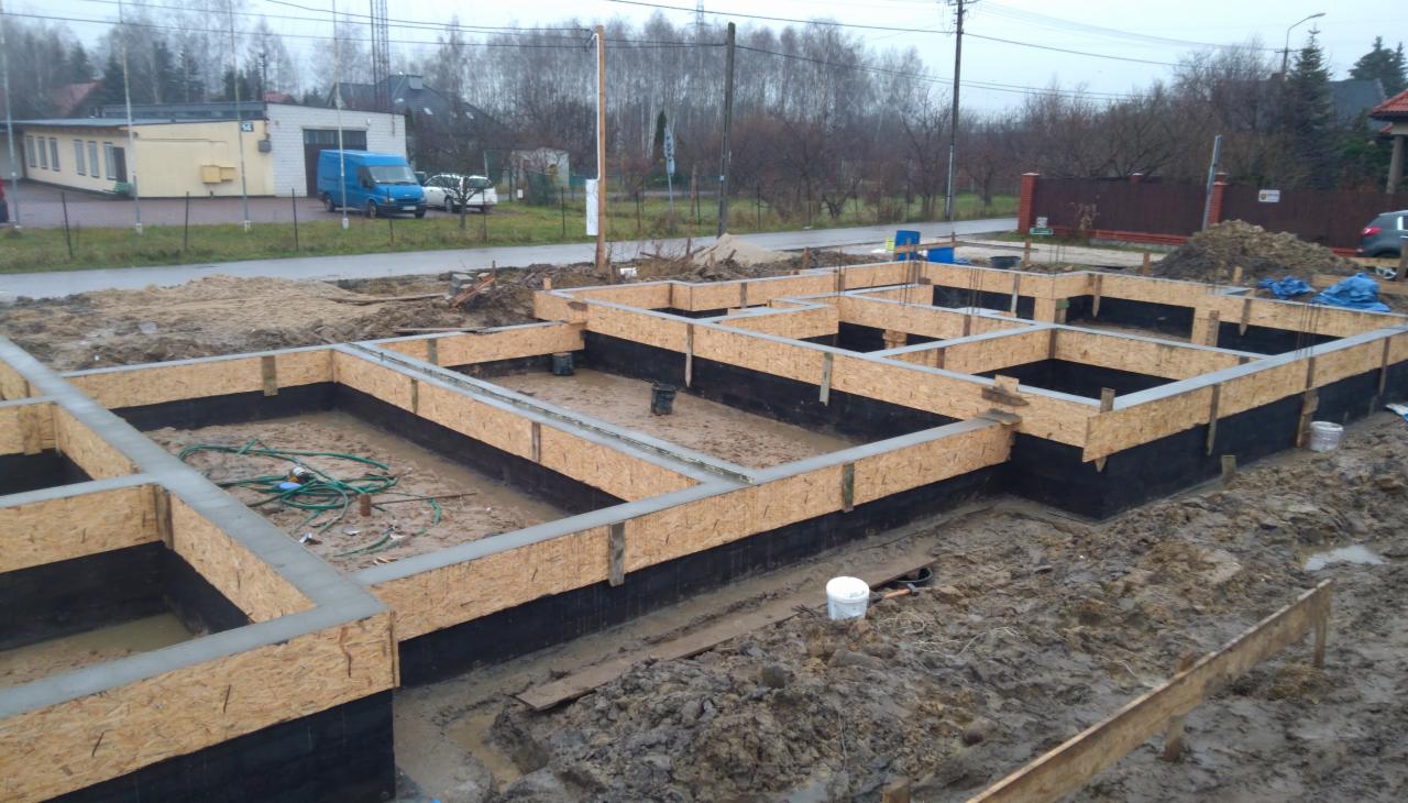 Wylane fundamenty z szalunkiem z płyty OSB i hydroizolacją pionową, widoczne zbrojenia i zalane betonem, na zewnątrz błoto i sprzęt budowlany.