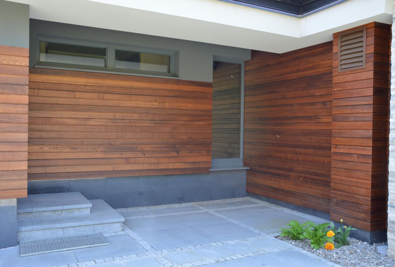 Nowoczesna elewacja domu wykończona poziomymi deskami z drewna w ciepłym odcieniu brązu, szare okna i schody wejściowe z kamienia, niewielka rabata z kwiatami u podstawy ściany.