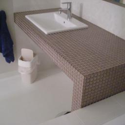 Virtuo Usługi Ogólnobudowlane Robert Matyjasik - Biała umywalka i blat z mozaiki w beżowych odcieniach, uzupełnione o wiszący, niebieski ręcznik i biały kosz na śmieci w jasnej łazience.