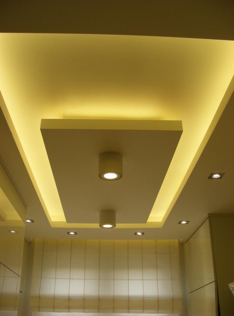 Nowoczesny sufit podwieszany z oświetleniem LED i cylindrycznymi lampami punktowymi. Kremowe ściany i meble w tle. Minimalistyczny design.