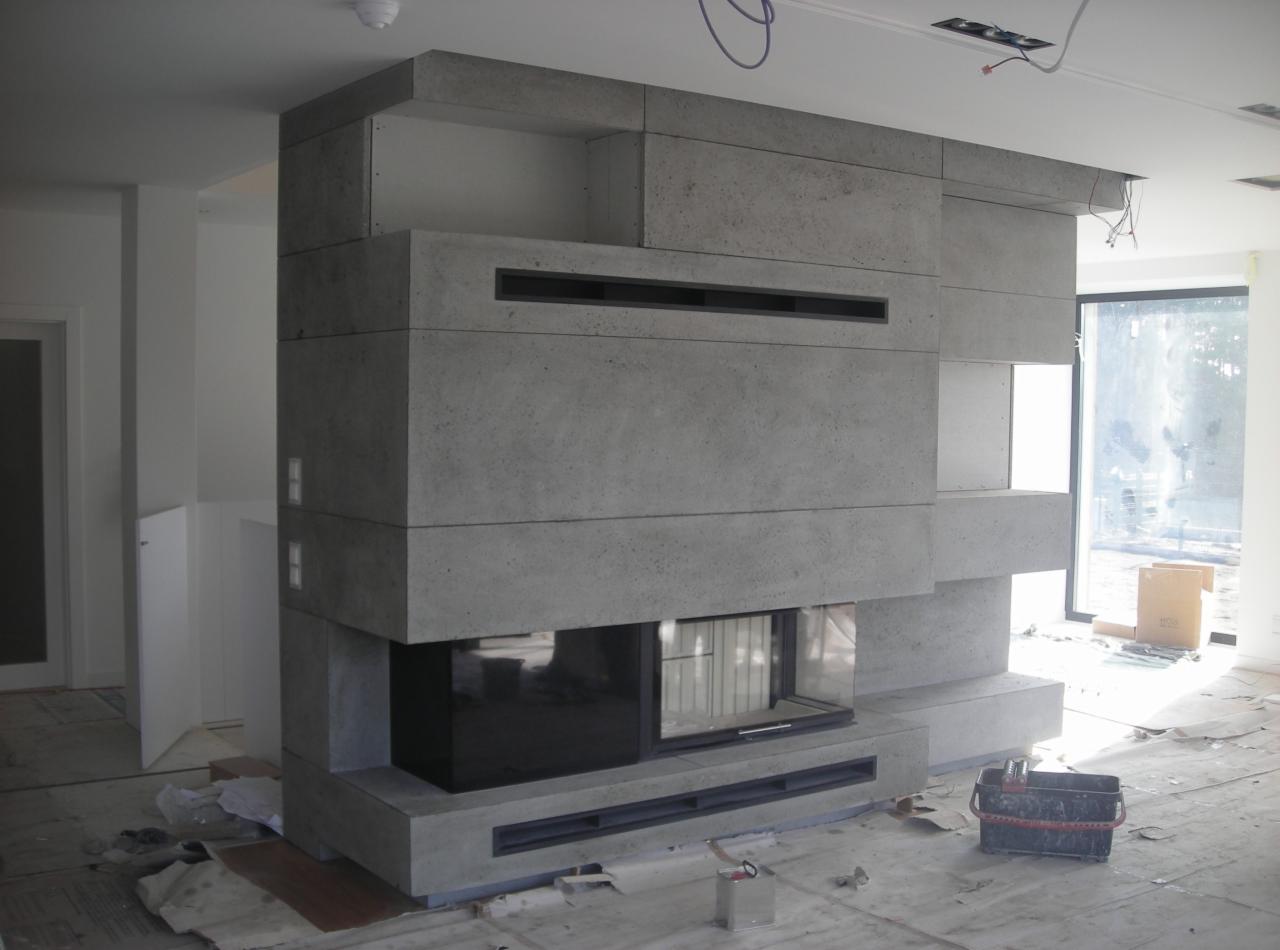 Nowoczesny kominek z betonu architektonicznego w surowym wnętrzu, widoczne niedokończone wykończenia podłogi i ściany, okno w tle.
