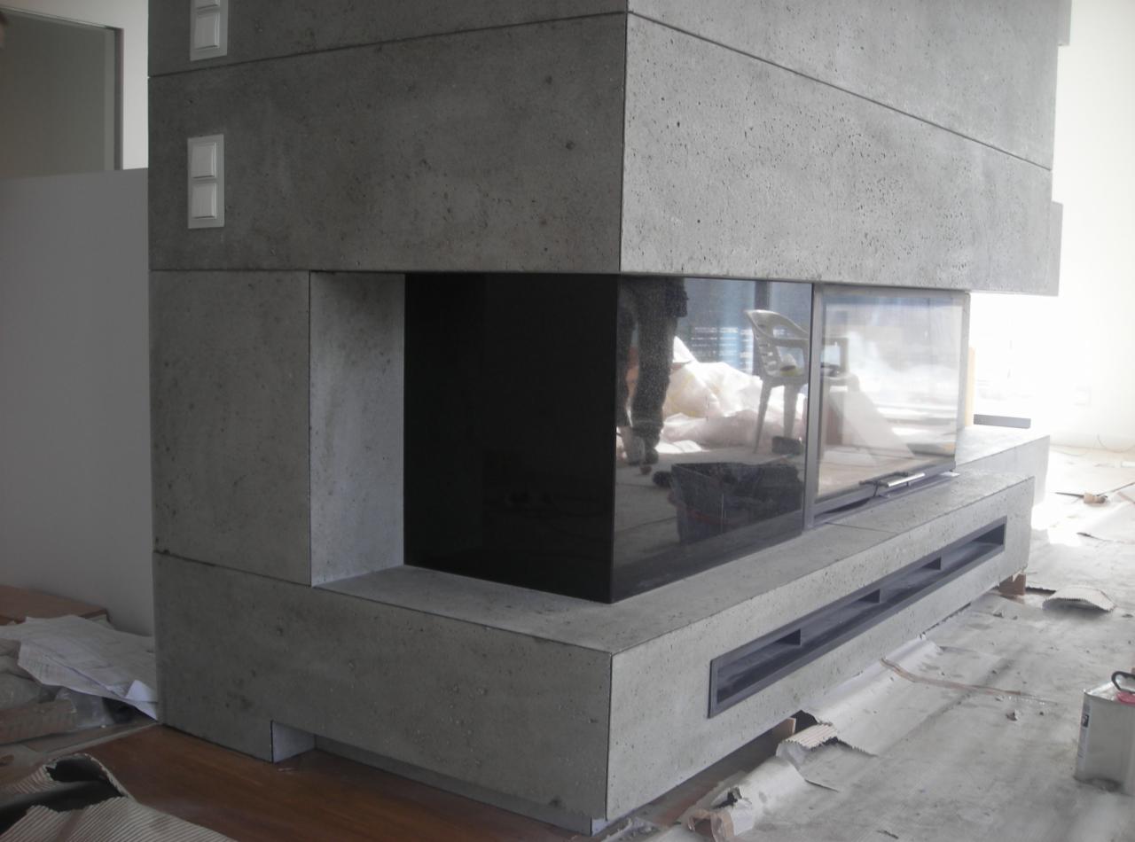 Betonowy kominek z czarnym szkłem, widoczne odbicie osoby, surowe wykończenie podłogi, nowoczesny design, jasne wnętrze, minimalistyczny styl.