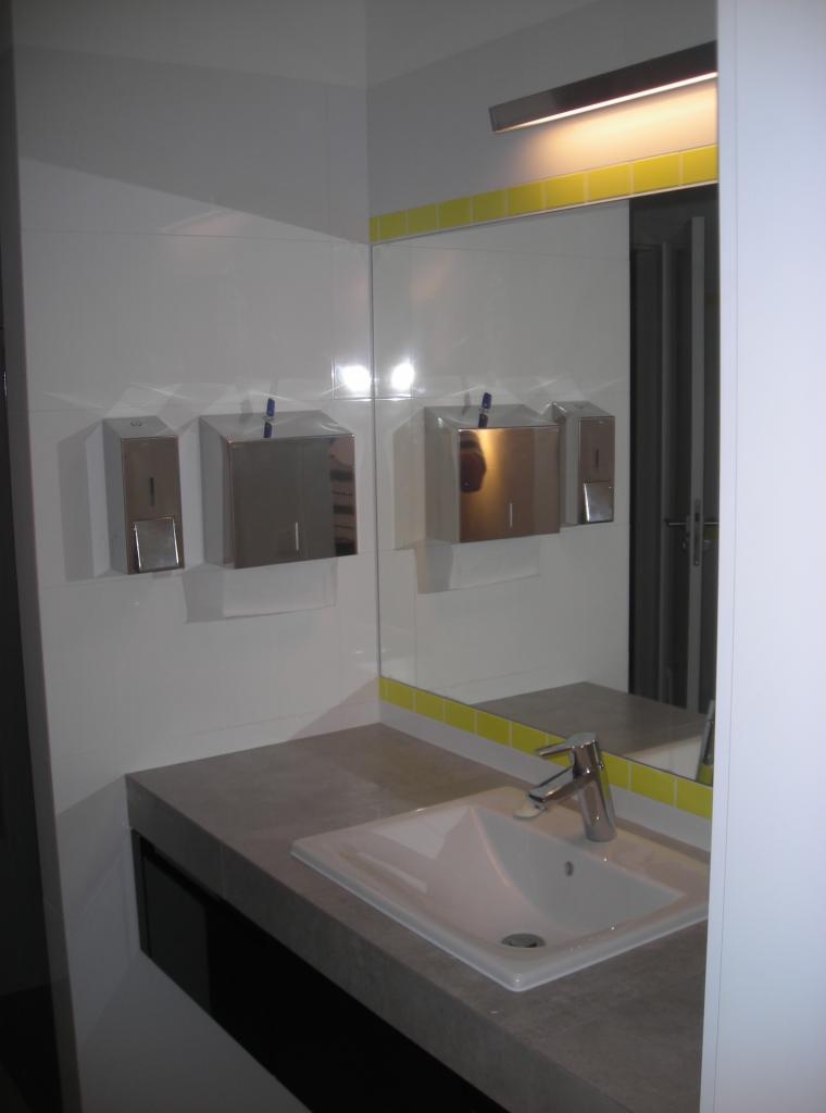 Nowoczesna łazienka z białą umywalką, szarym blatem i lustrem z żółtą obwódką. Na ścianie dozowniki mydła i ręczników ze stali nierdzewnej.