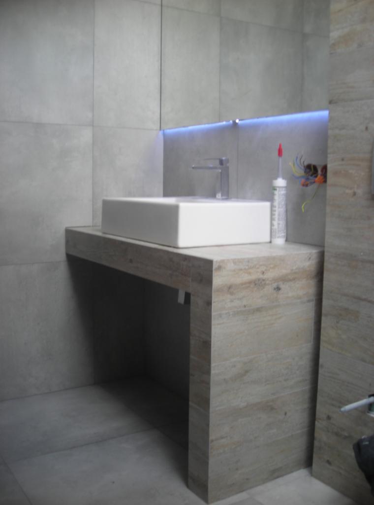 Nowoczesna łazienka: biała umywalka na blacie z płytek imitujących drewno, podświetlane lustro, szare płytki ścienne i podłogowe. Widoczne kable i tuba z silikonem.