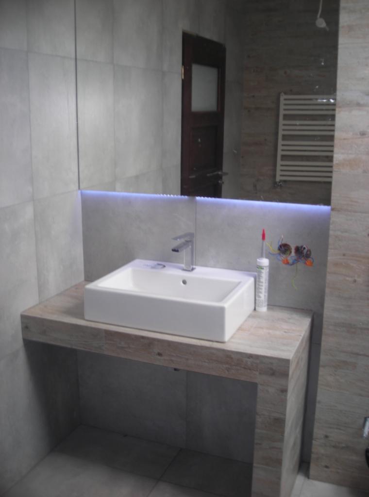 Nowoczesna umywalka nablatowa z baterią, podświetlana LED, na blacie z imitacją drewna. W tle lustro z widoczną grzejnikiem i fragmentem drzwi.