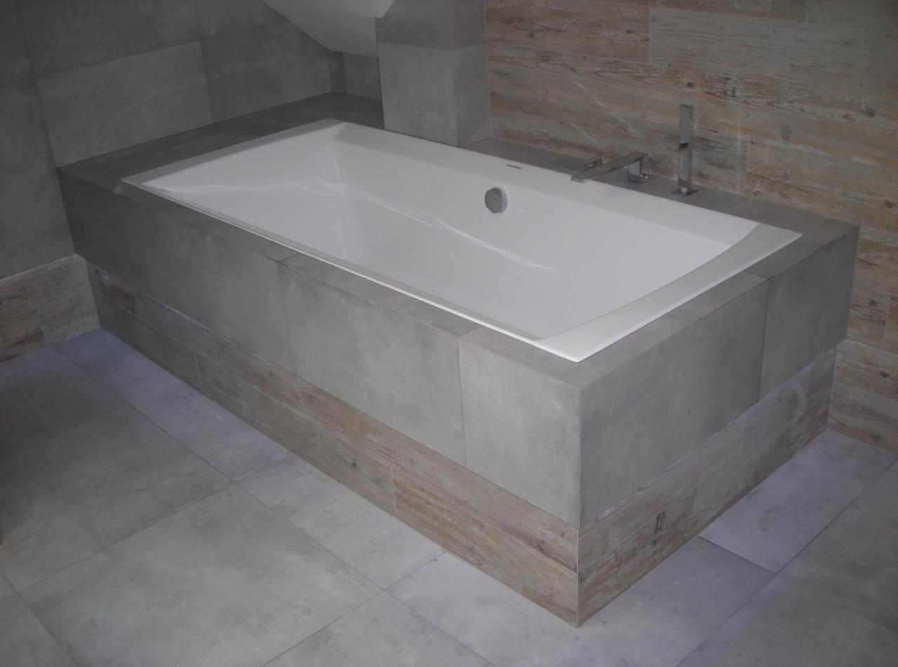 Nowoczesna wanna obudowana szarymi płytkami i drewnopodobnym dekorem w łazience z szarymi płytkami. Bateria wannowa chromowana.