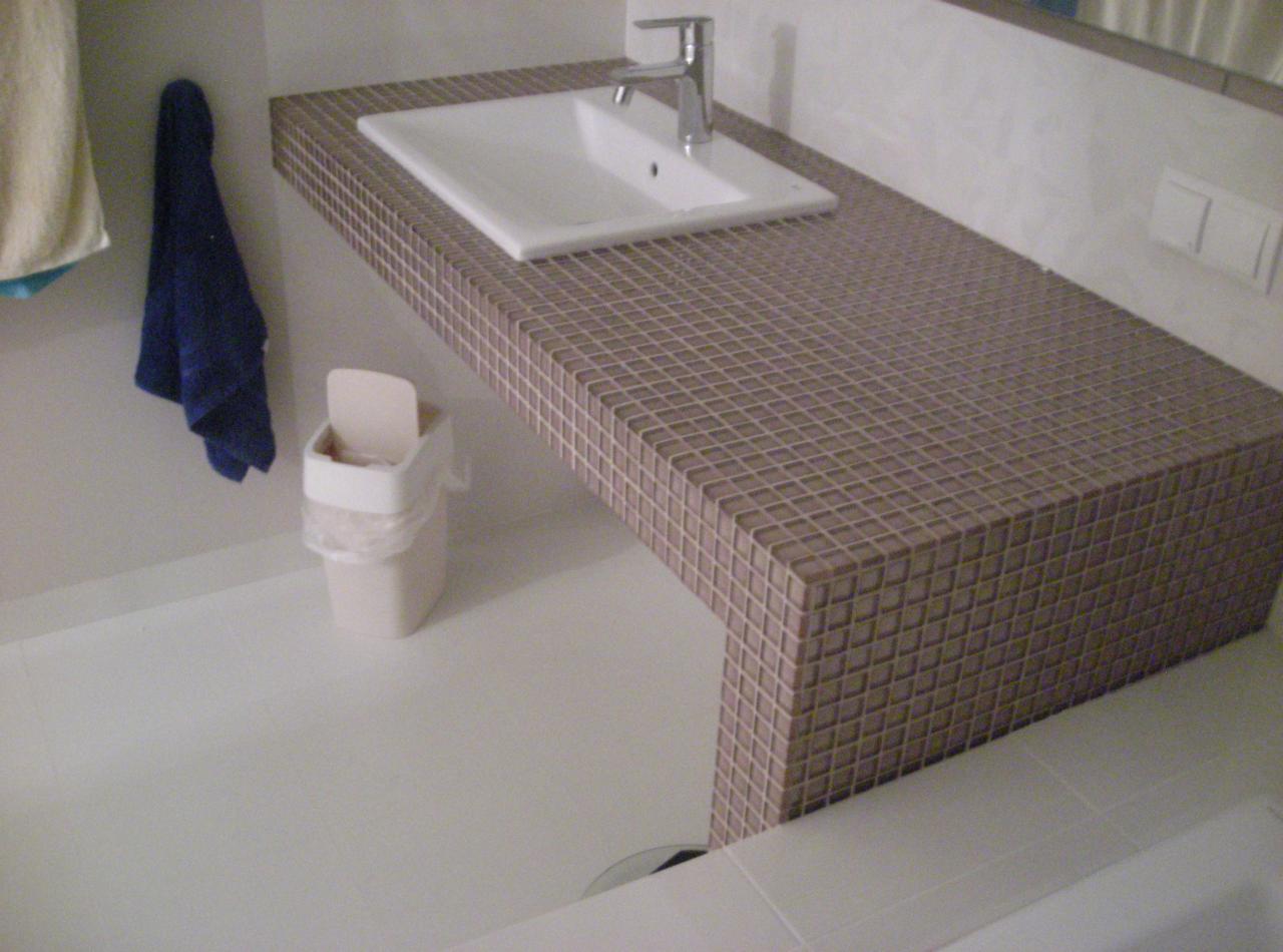 Biała umywalka i blat z mozaiki w beżowych odcieniach, uzupełnione o wiszący, niebieski ręcznik i biały kosz na śmieci w jasnej łazience.