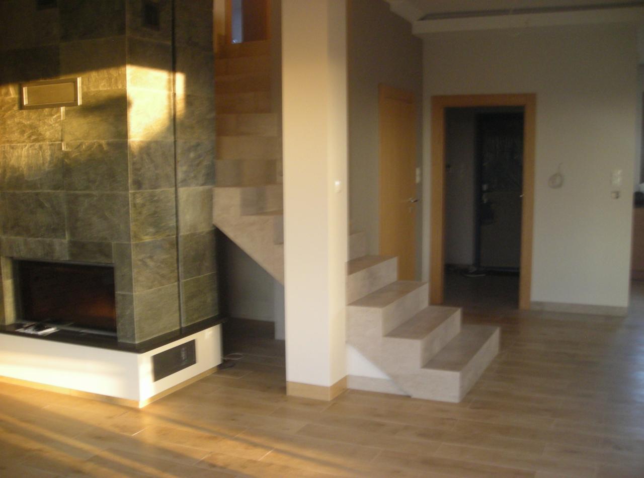 Salon po remoncie: kamienny kominek z wkładem, drewnopodobna podłoga, schody z jasnego kamienia, drewniane drzwi i wykończone wejście do pokoju.