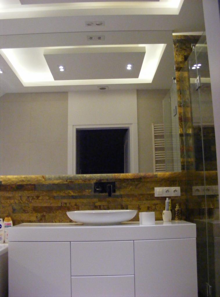 Nowoczesna łazienka z białymi szafkami, umywalką i lustrem. Ściana z dekoracyjną okładziną. Podwieszany sufit z oświetleniem punktowym.