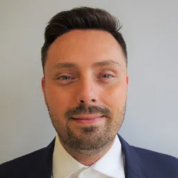 Portret mężczyzny w garniturze z delikatnym zarostem na tle jasnoszarej ściany.