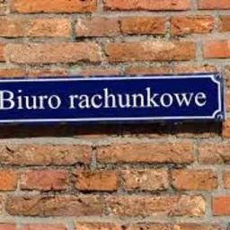 Niebieska tabliczka z białym napisem 'Biuro rachunkowe' umieszczona na ceglanej ścianie, widoczna z bliska, z wyraźną fakturą cegieł i detalami tabliczki.