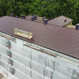 Widok z góry na budynek z brązowym dachem w trakcie prac remontowych, zabezpieczony rusztowaniami i siatką z logo farb Caparol. Na dachu widoczne kominy, a w tle zielone korony drzew.