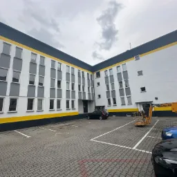 Wielopiętrowy budynek biurowy po remoncie elewacji w kolorach białym, szarym, czarnym i żółtym, z podnośnikiem koszowym Haulotte HA16F na parkingu przed budynkiem.