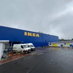 Duży, niebieski budynek IKEA z żółtym logo, dwa białe busy zaparkowane przed wejściem, pracownicy w kamizelkach odblaskowych i kaskach, pochmurne niebo.