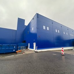 RS-MAL - Niebieski budynek magazynowy z widocznymi oknami, rampą załadunkową i kontenerem na śmieci, sfotografowany w pochmurny dzień z poziomu gruntu.