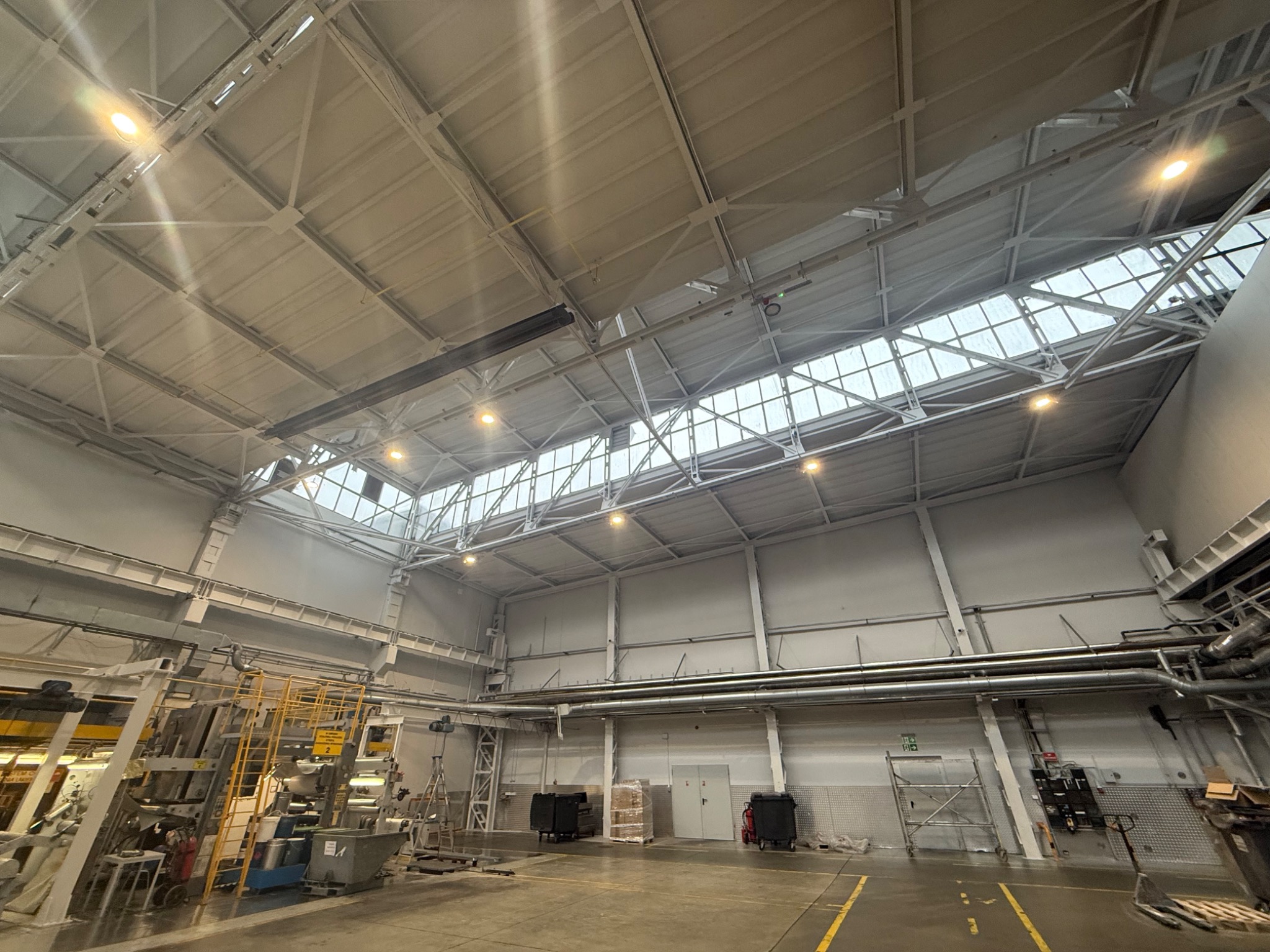 Wnętrze hali produkcyjnej z widoczną konstrukcją dachu, oświetleniem i instalacjami, ściany pomalowane na jasny kolor.