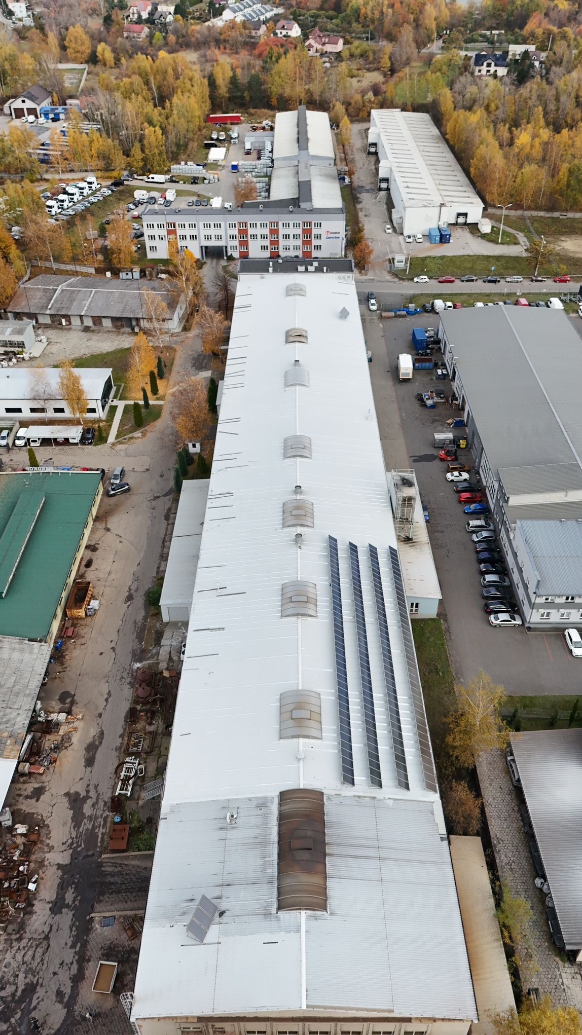 Widok z góry na długi, biały dach hali przemysłowej z rzędami paneli słonecznych i świetlikami, otoczonej jesiennymi drzewami i budynkami.