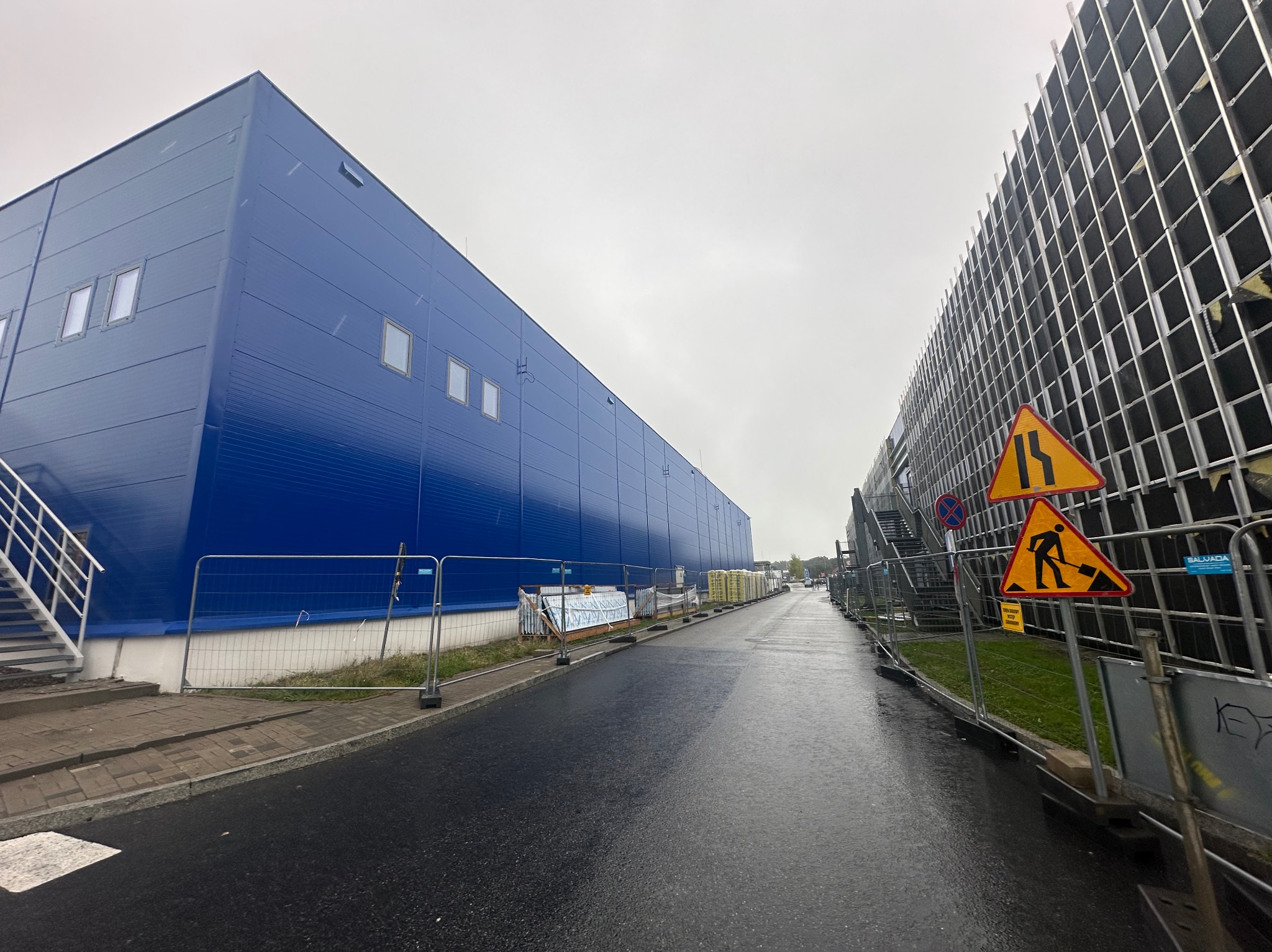 Nowoczesny budynek przemysłowy z niebieską elewacją panelową i zewnętrznymi schodami ewakuacyjnymi, widoczny z poziomu mokrej, asfaltowej drogi w trakcie budowy lub remontu, z tymczasowymi...