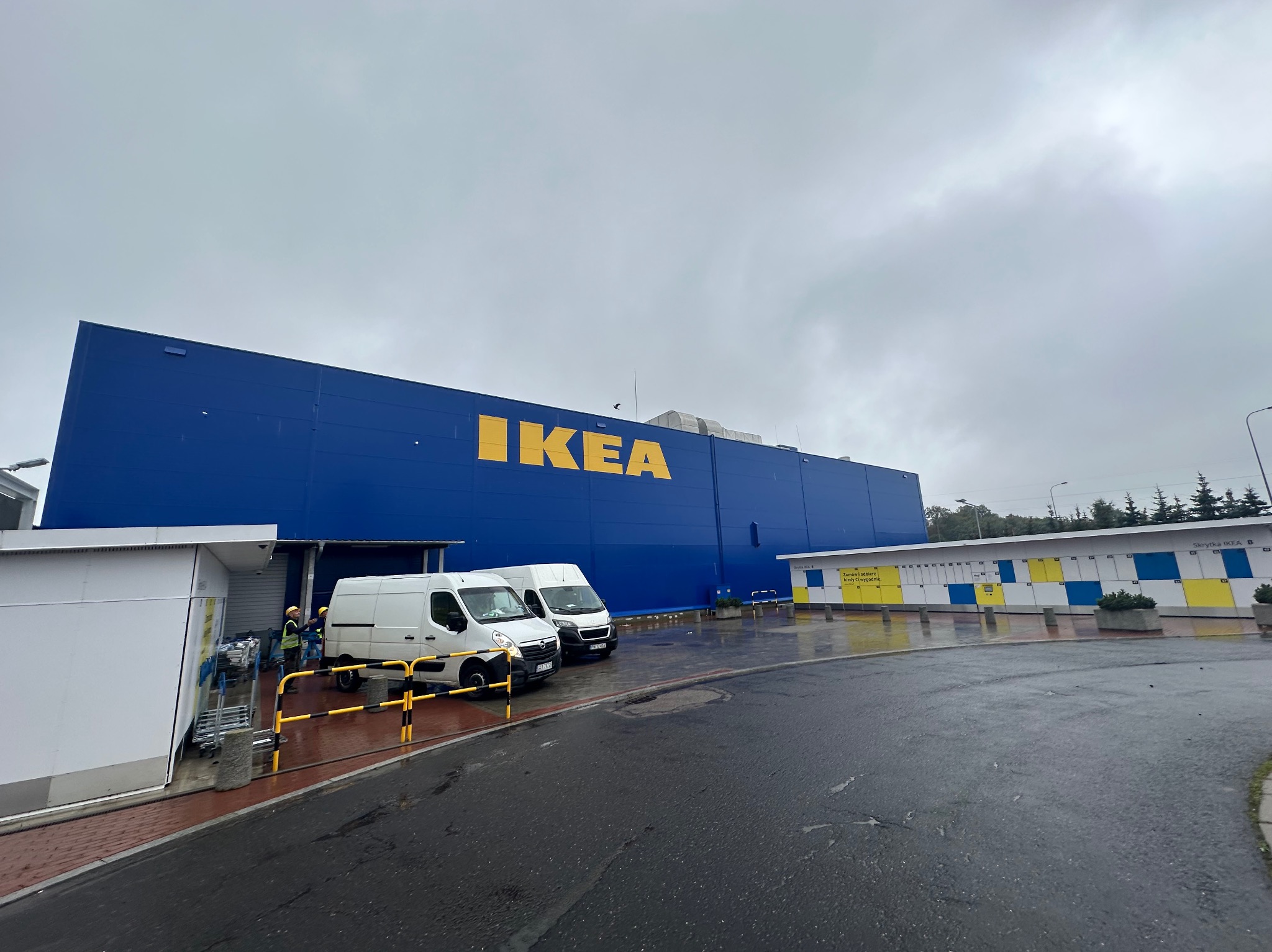 Duży, niebieski budynek IKEA z żółtym logo, dwa białe busy zaparkowane przed wejściem, pracownicy w kamizelkach odblaskowych i kaskach, pochmurne niebo.
