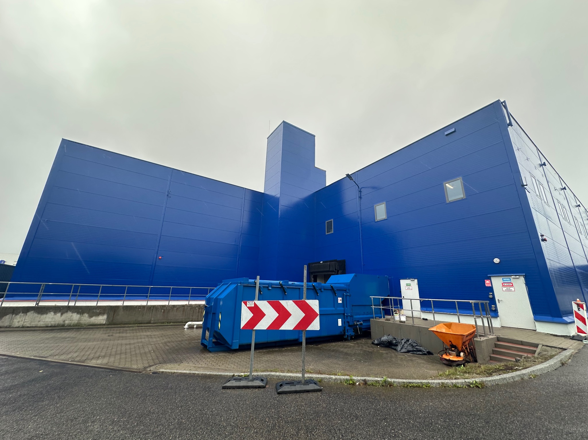 Niebieski budynek przemysłowy z kontenerem na śmieci i rozsiewaczem soli na zewnątrz, widok z dołu w pochmurny dzień.