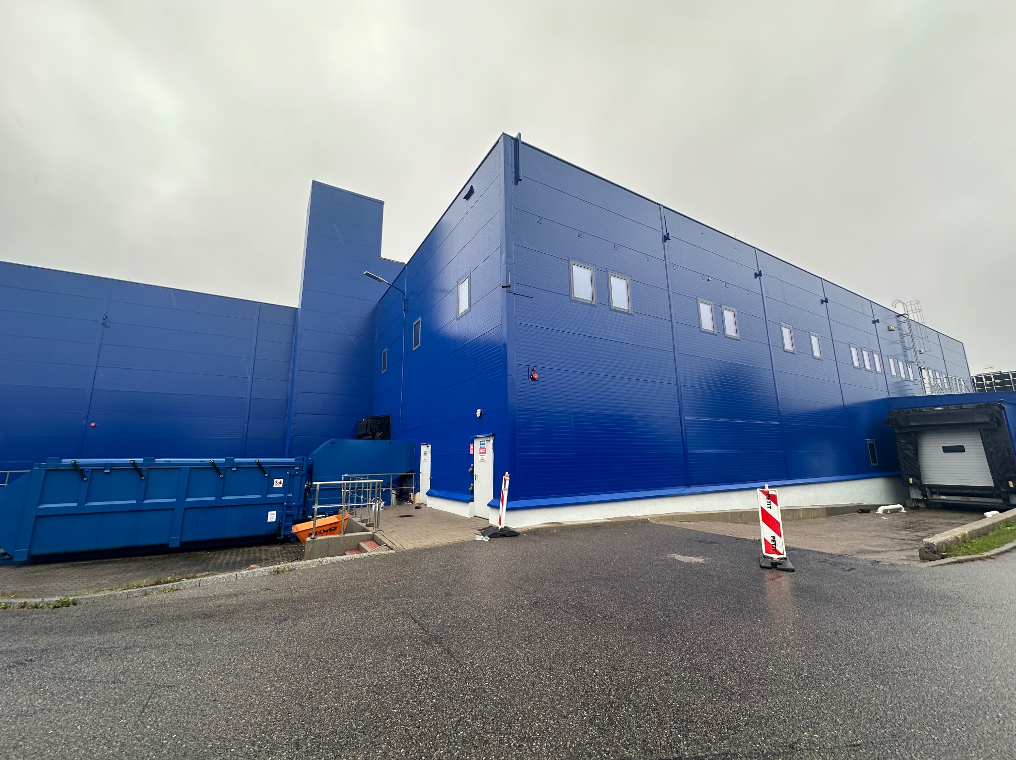 Niebieski budynek magazynowy z widocznymi oknami, rampą załadunkową i kontenerem na śmieci, sfotografowany w pochmurny dzień z poziomu gruntu.