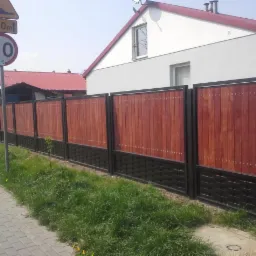 Nowoczesne ogrodzenie panelowe z drewnianym wypełnieniem w odcieniach brązu i czarną, plecioną podstawą, widoczne z poziomu chodnika, z domem w tle.