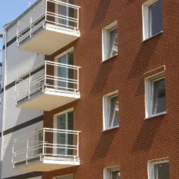 Elewacja budynku z cegły z białymi balkonami i balustradami. Widoczne rusztowania z lewej strony budynku i okna z białymi ramami.