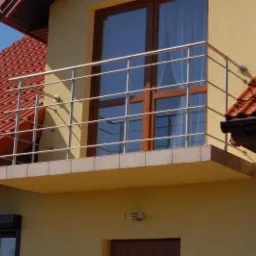 Balkon z nierdzewną balustradą na tle żółtej elewacji domu z czerwoną dachówką, widok z dołu. Balustrada z poziomymi prętami, brązowe ramy okien.