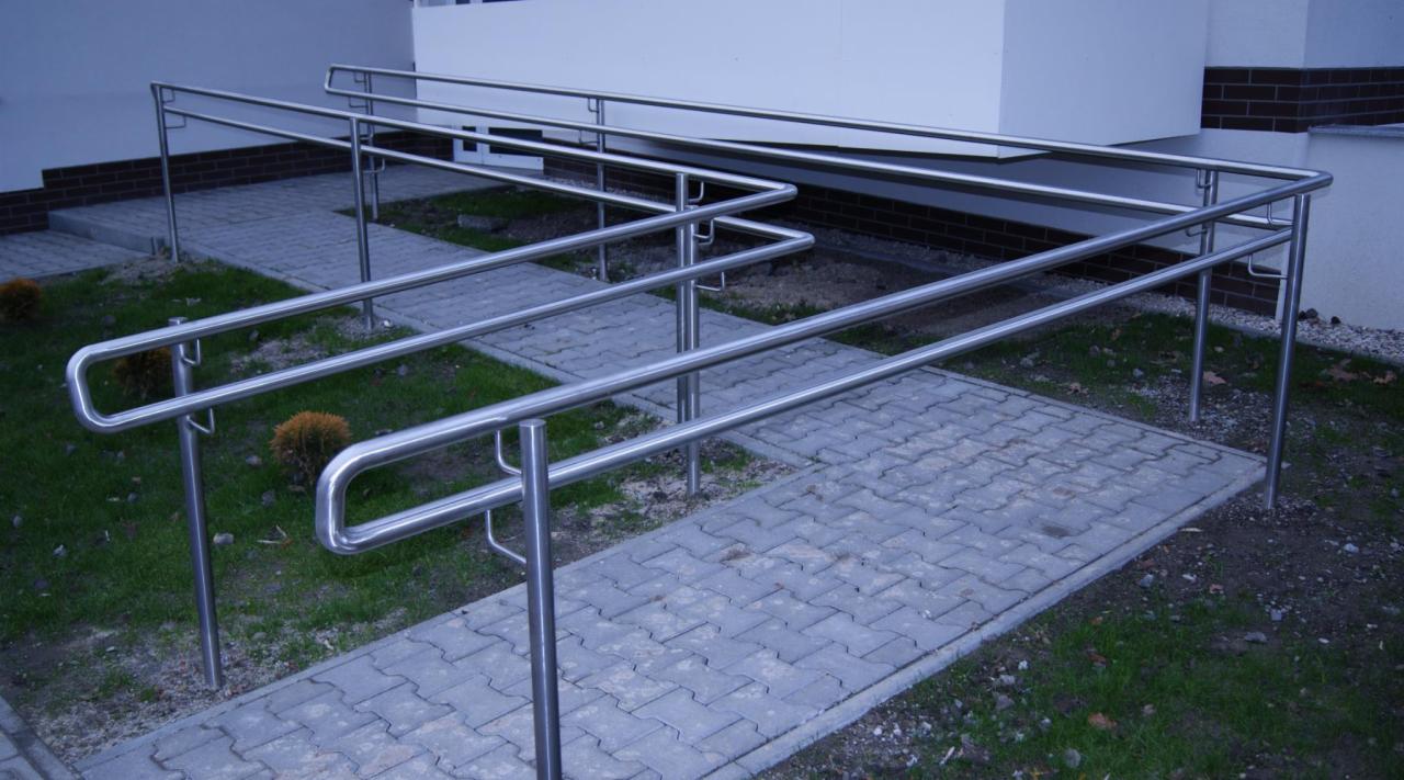 Zewnętrzne, metalowe poręcze ze stali nierdzewnej, ułatwiające dostęp do budynku. Szara kostka brukowa, zielona trawa i fragment elewacji w tle.
